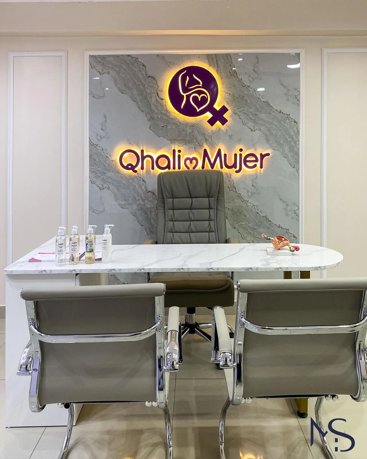 Consultorio con escritorio de mármol blanco, sillas tapizadas y muro de porcelanato efecto mármol con logotipo en neón retroiluminado