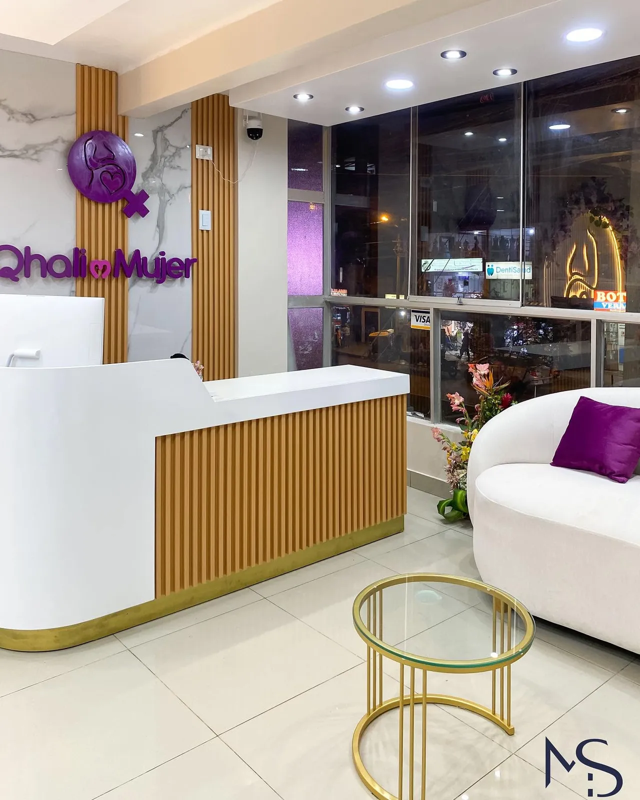 Recepción de la clínica Qhali Mujer con mostrador blanco, frente de listones de madera, logotipo iluminado y sofá curvo blanco