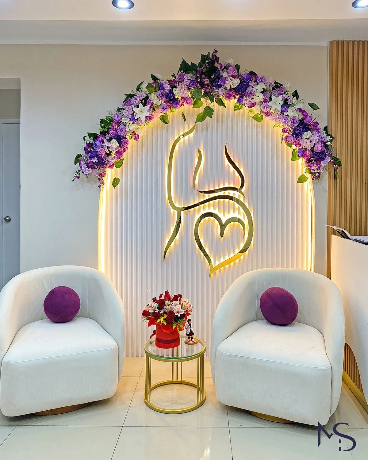 Sala de espera con dos butacas curvas blancas, mesa auxiliar dorada y arco floral retroiluminado con escultura en dorado