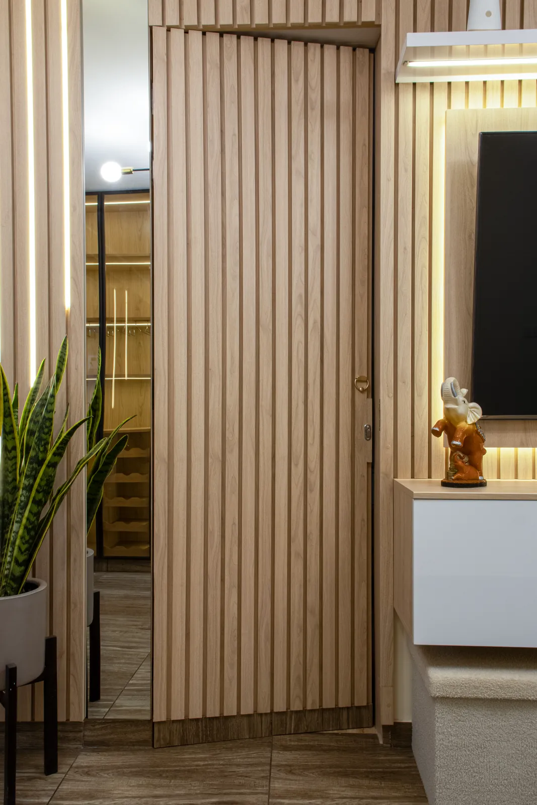 Puerta oculta integrada en un panel de listones de roble, junto a un espejo retroiluminado y una sansevieria
