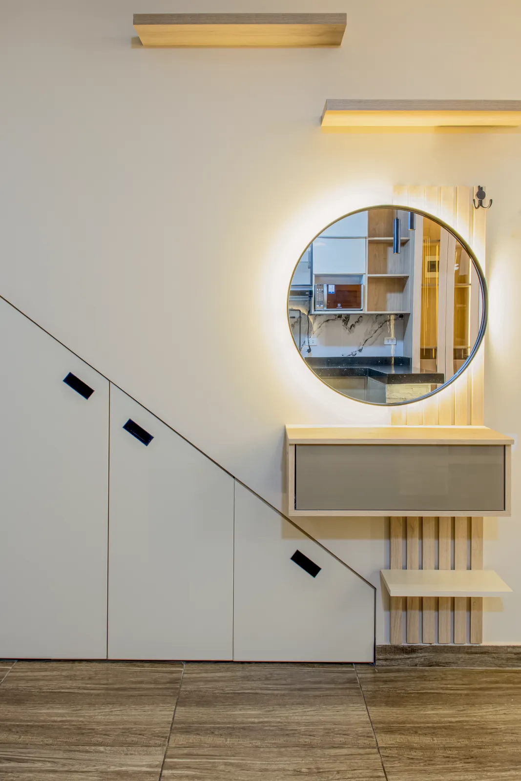 El mismo recibidor bajo escalera con el espejo circular retroiluminado encendido, bañando de luz cálida los listones — Santiago de Surco